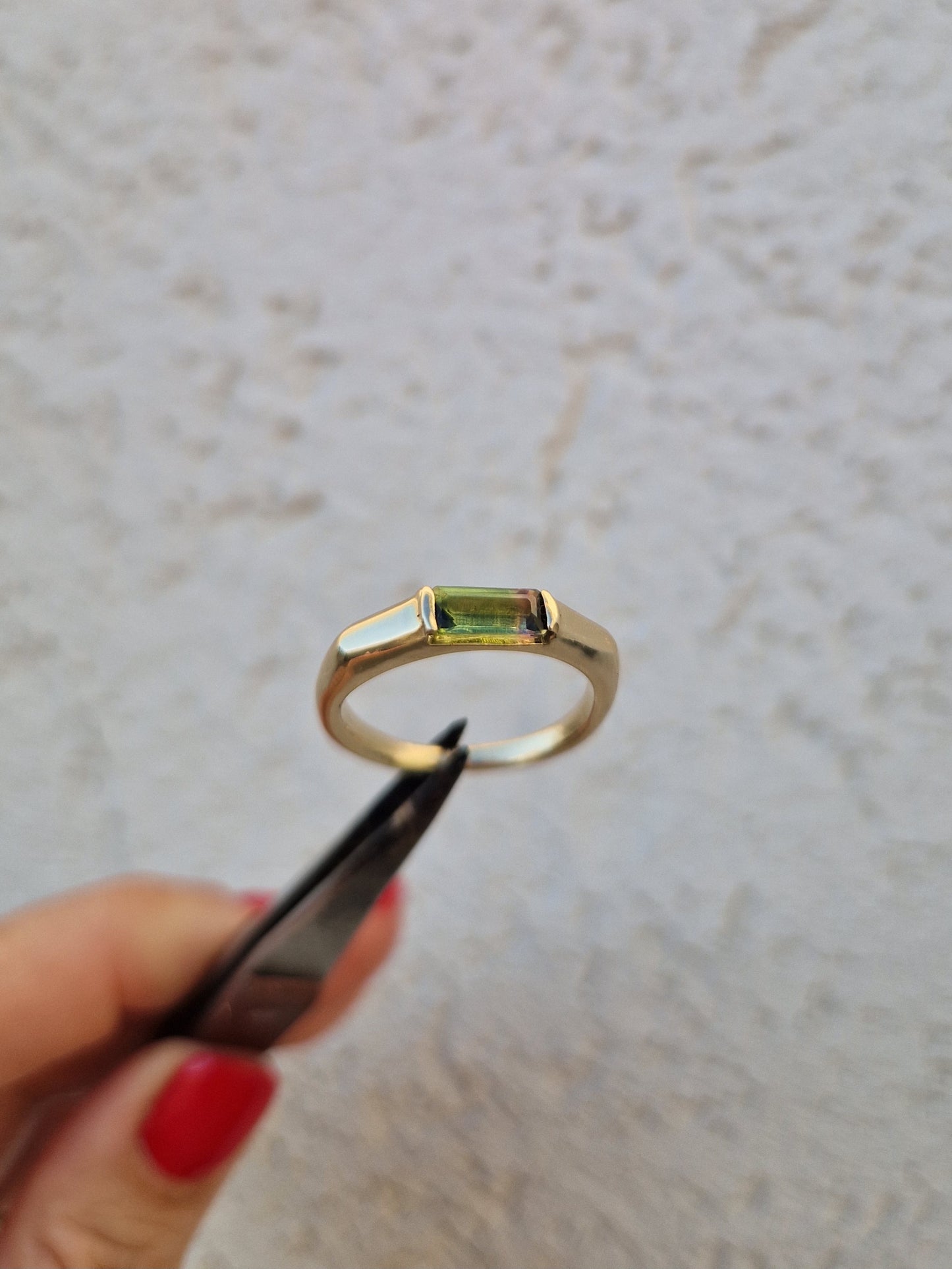 A gold ring with a rectangular green gemstone held by tweezers against a textured off-white wall background.